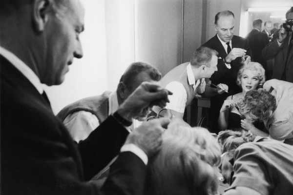 A black and white photo of Marilyn Monroe receiving makeup and hair preparation on the set of movie Let's Make Love 1960.
